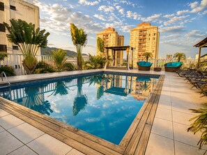 A rooftop pool with a wooden deck, lounge chairs, and a view of surrounding buildings. - Don Marco's Hotel (Guarujá)