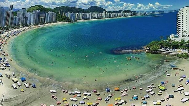 A coastal beach with a wide sandy area, clear blue waters, and a row of high-rise buildings in the background. - Don Marco's Hotel (Guarujá)