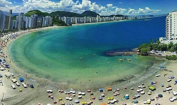 A coastal beach with a wide sandy area, clear blue waters, and a row of high-rise buildings in the background. - Don Marco's Hotel (Guarujá)