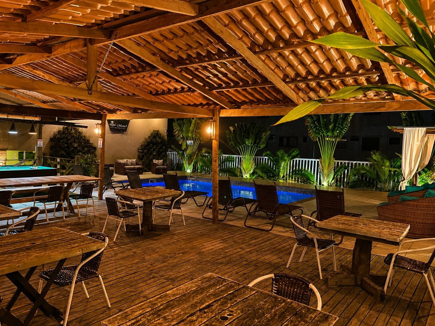 Zona de piscina exterior con mesas y sillas de madera, rodeada de plantas y un pérgola de madera.