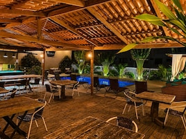 An outdoor pool area with wooden tables and chairs, surrounded by plants and a wooden pergola.