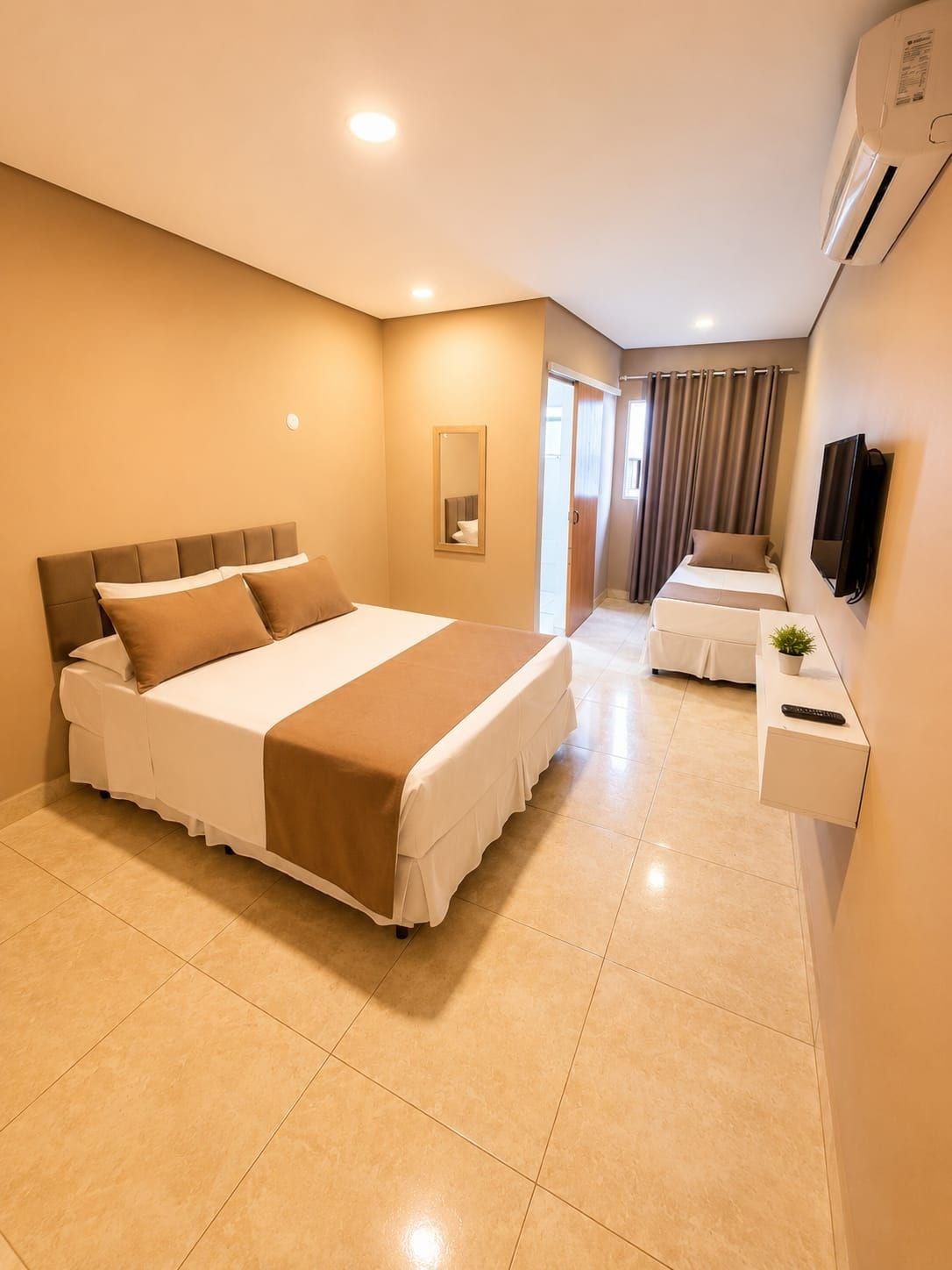 A hotel room with a large bed, a bedside table with a plant, a TV mounted on the wall, and an air conditioning unit.