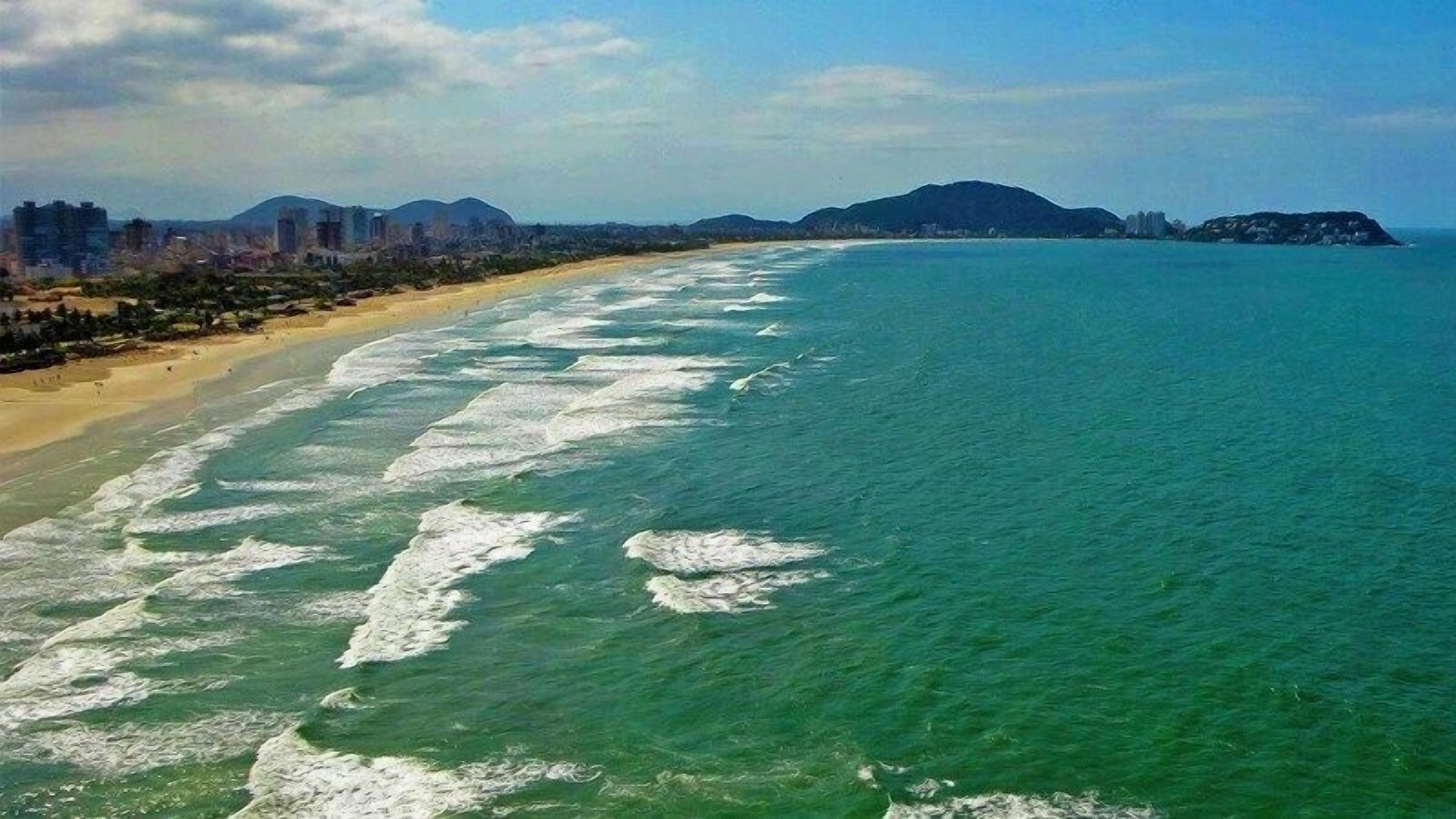 Une vue aérienne d’une plage aux eaux cristallines, bordée d’un rivage sablonneux et surplombée de collines lointaines.