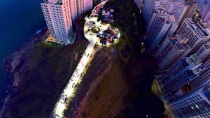 An aerial view of a coastal cityscape with high-rise buildings, a winding road, and a prominent hill with a circular structure. - Don Marco's Hotel (Guarujá)