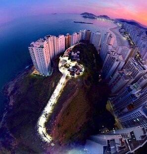 An aerial view of a coastal cityscape with high-rise buildings, a winding road, and a prominent hill with a circular structure. - Don Marco's Hotel (Guarujá)