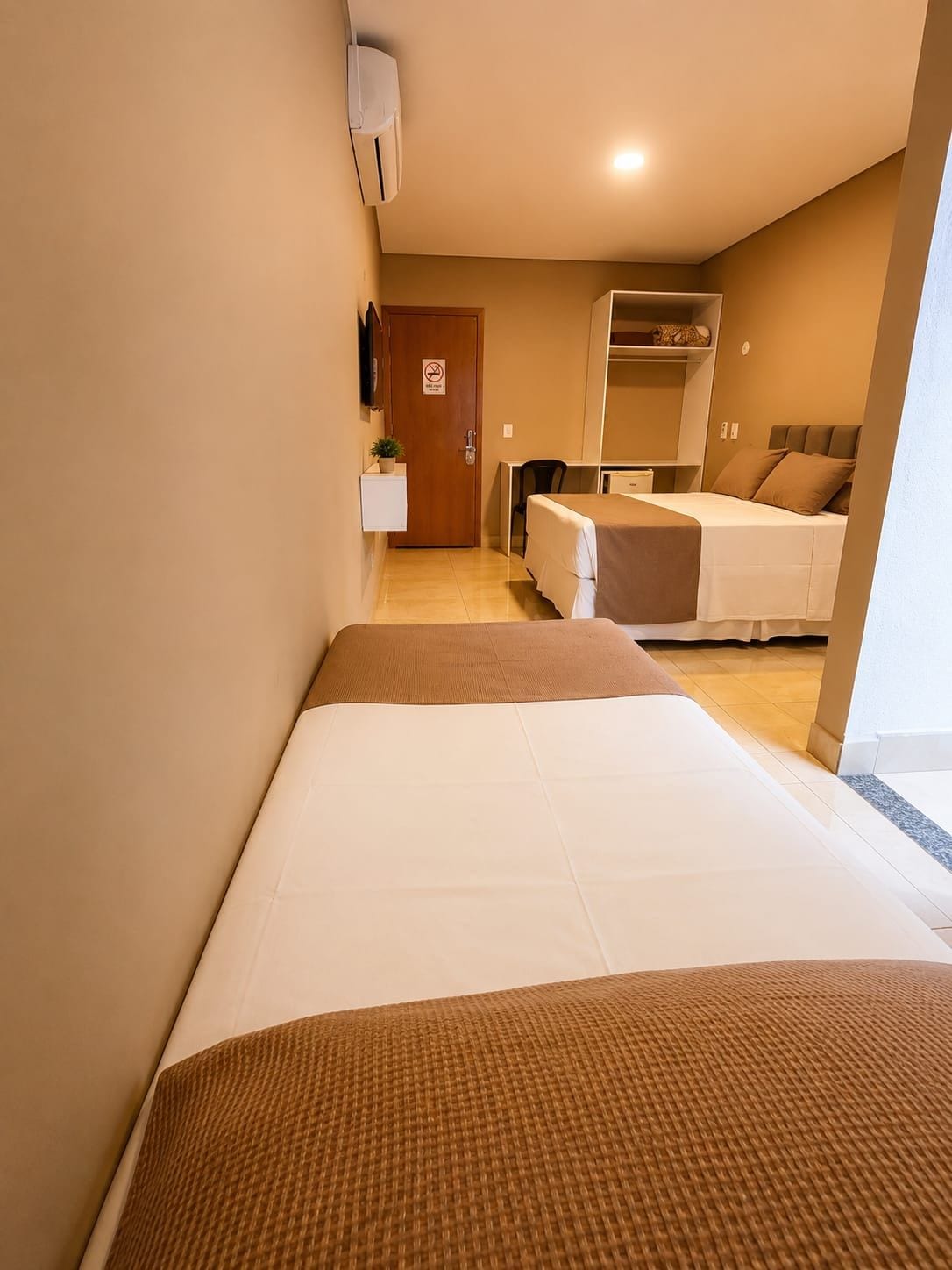 Ett hotellrum med en säng, ett skrivbord och en stol. Det finns en dörr med en skylt på, och en växt på en hylla.