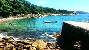 A coastal scene with clear water, rocky shore, and a boat. - Don Marco's Hotel (Guarujá)