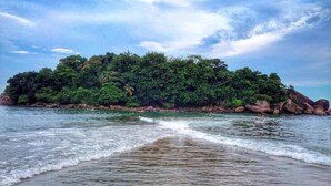 A beach with waves gently lapping at the shore, a small island with dense vegetation in the background, and a clear sky above. - Don Marco's Hotel (Guarujá)