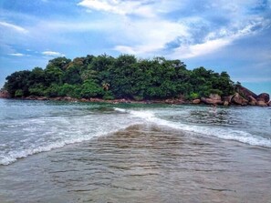 A beach with waves gently lapping at the shore, a small island with dense vegetation in the background, and a clear sky above. - Don Marco's Hotel (Guarujá)