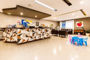 Un área de recepción con mostrador de diseño geométrico, un comedor con mesa y sillas, y una sala de estar con sofá y una planta en maceta grande.