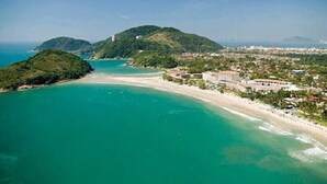 A coastal area with a beach, a small hill with a building, and a town in the background. - Don Marco's Hotel (Guarujá)