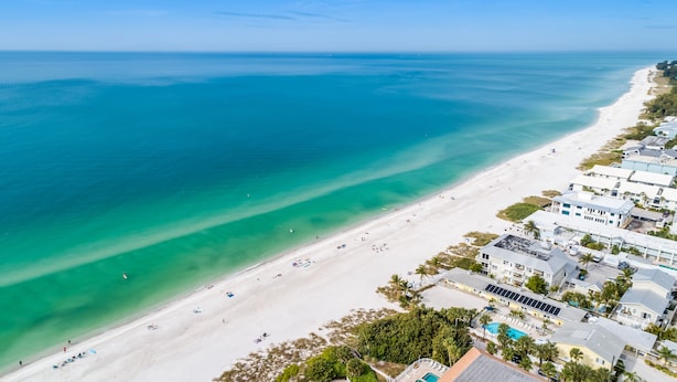 An aerial view of a beach with a clear turquoise sea, a sandy shore, and a row of white buildings with a swimming pool. - Tropical Anna Maria Island Villa (Holmes Beach)