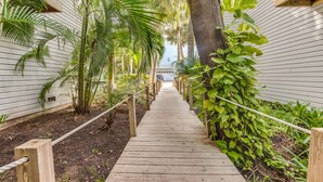 A wooden pathway flanked by lush greenery and white siding buildings. - Tropical Anna Maria Island Villa (Holmes Beach)