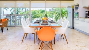 Uma sala de jantar moderna com uma mesa grande posta para seis pessoas, rodeada por cadeiras brancas, uma fruteira e vista para vegetação exuberante através de grandes janelas.