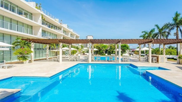 Piscina com pergolado, cadeiras de descanso e um edifício moderno com varandas.