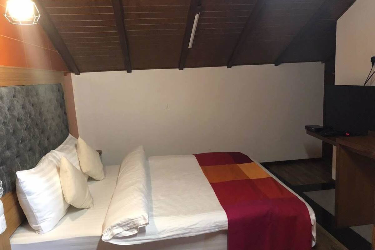 Un lit simple avec une literie blanche et une couverture rouge, une tête de lit en bois et une petite table de chevet en bois sur laquelle se trouve un téléviseur.
