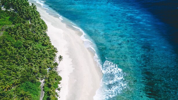 An aerial view of a beach with clear turquoise waters and white sand, surrounded by dense green vegetation. - Millennium Island Residence (Fuvahmulah)