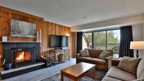 A cozy living room with a fireplace, a sofa set, a television, and a view of the outdoors. - Charming, Renovated 2BR/2BA Mt Green Condo: Steps to Killington 3C13/A (Killington)
