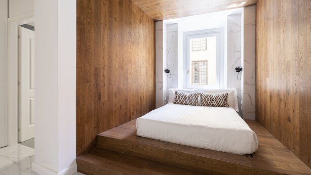 A bedroom with a wooden floor, a bed with white bedding, and a view of a building through an open door. - Via del Tritone (Rome)