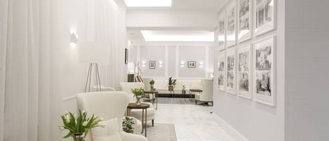 A modern living room with a white sofa, a round table with a vase of flowers, and a coffee table. There are framed pictures on the wall and a staircase in the background.