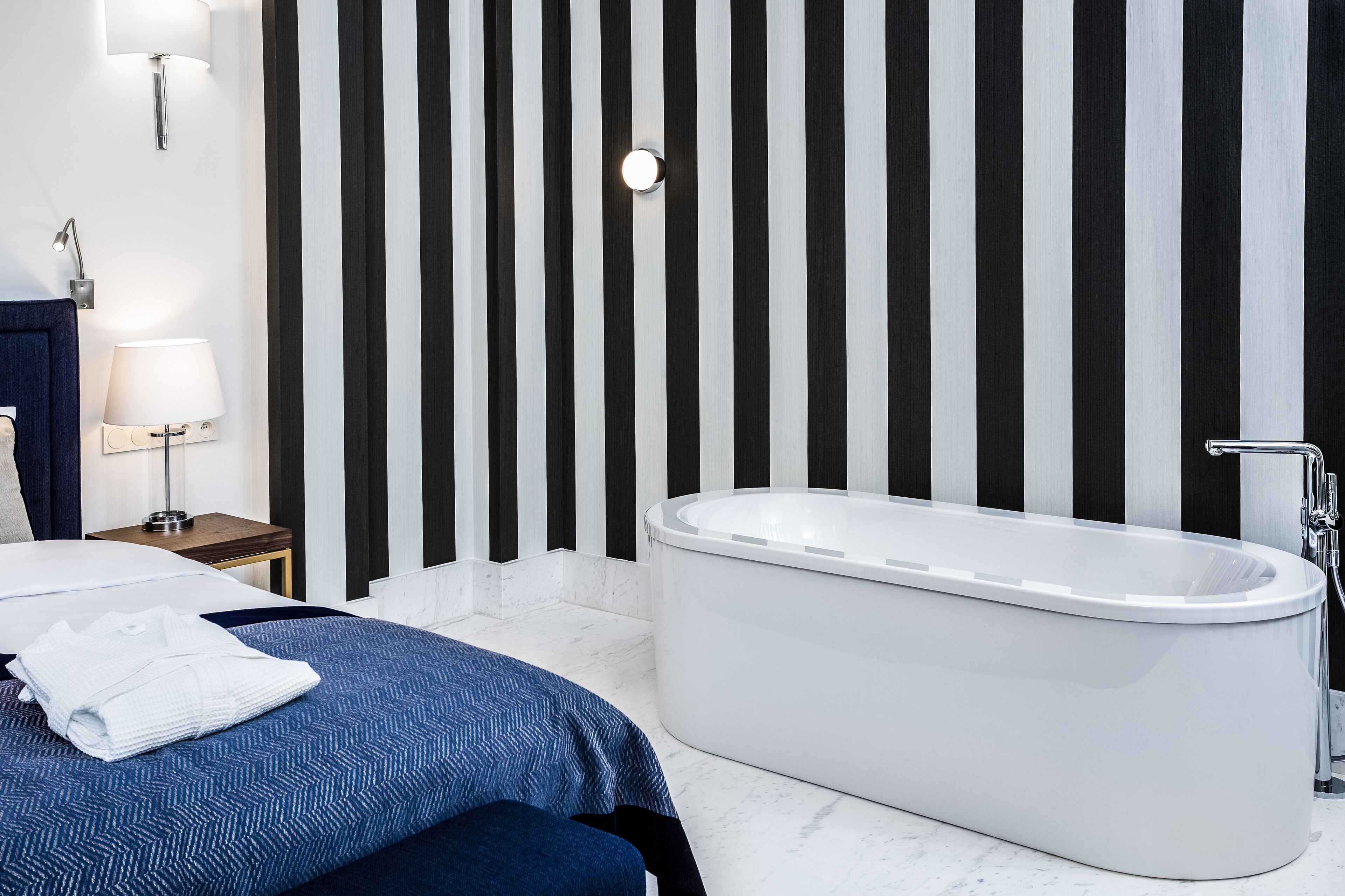 A modern bathroom with a freestanding bathtub, a bedside table with a lamp, and a striped wall.