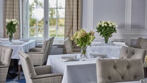 Un comedor con mesas dispuestas para dos, cada una con un florero, sillas y vistas al exterior a través de amplios ventanales.