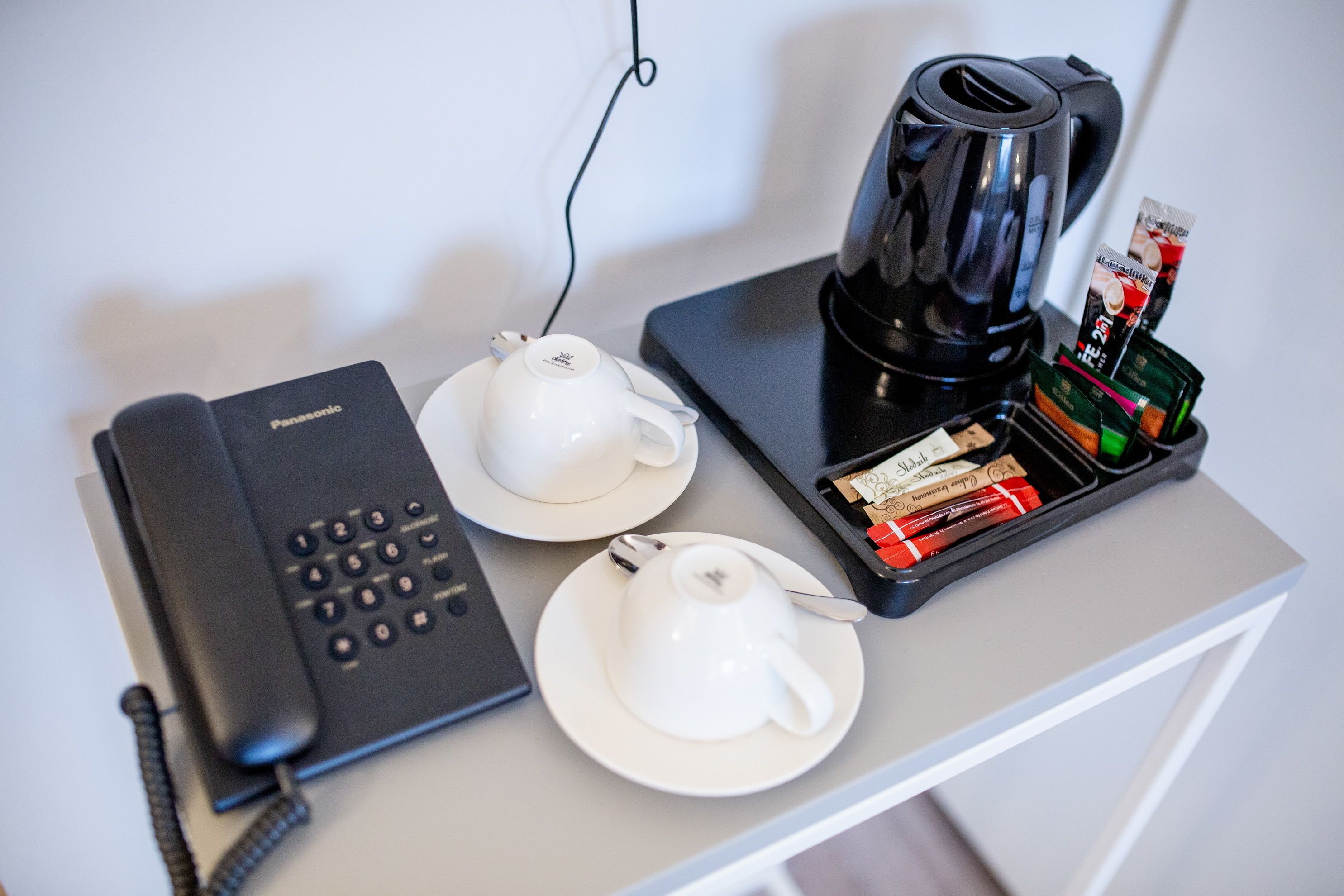 A hotel room with a telephone, two white teacups on a tray, a black coffee maker, and a tray of assorted snacks.