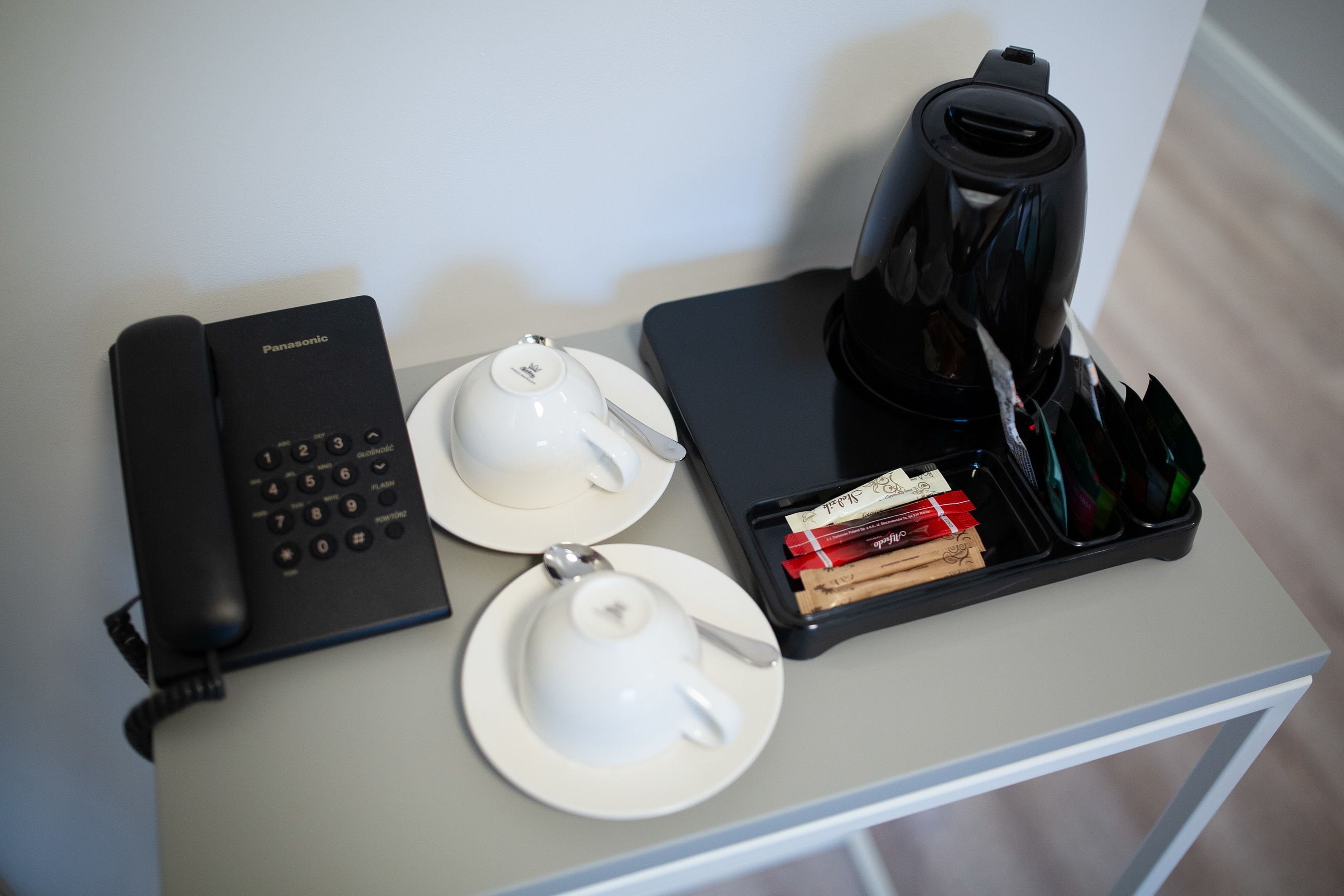 Een tafel met een zwart Panasonic telefoon, twee witte theekopjes, een zwarte koffiezetapparaat, en een dienblad met boeken en een zakdoos.