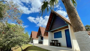 A row of small, single-story buildings with a red-tiled roof and a blue door, surrounded by greenery and palm trees. - Tijota Park Hotel Fazenda (Santana Do Paraiso)