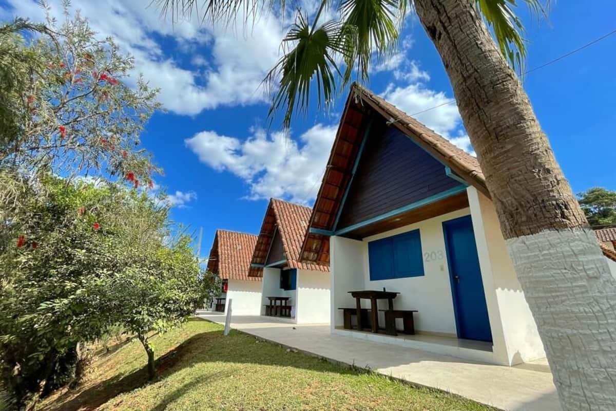 A row of small, single-story buildings with a red-tiled roof and a blue door, surrounded by greenery and palm trees.