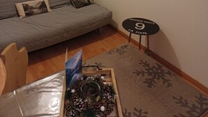 Una sala de estar acogedora con un sofá gris, un televisor montado, una pequeña mesa de madera con un objeto decorativo y una ventana con persianas.