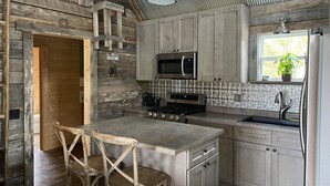 A rustic kitchen with wooden walls, a stone accent wall, a built-in stove, a microwave, and a sink with a window above it.