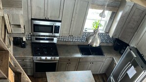 A modern kitchen with a stainless steel refrigerator, oven, and microwave. There is a wooden staircase leading to a seating area with a leather sofa and two chairs.