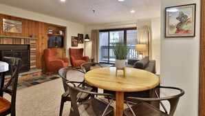 A cozy living room with a fireplace, a dining area with a round table, and a large window with curtains. - Snowplace Like Home at Mountain Green 1BR/1BA (Killington)