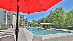 A swimming pool area with lounge chairs and umbrellas, surrounded by buildings and trees. - Snowplace Like Home at Mountain Green 1BR/1BA (Killington)