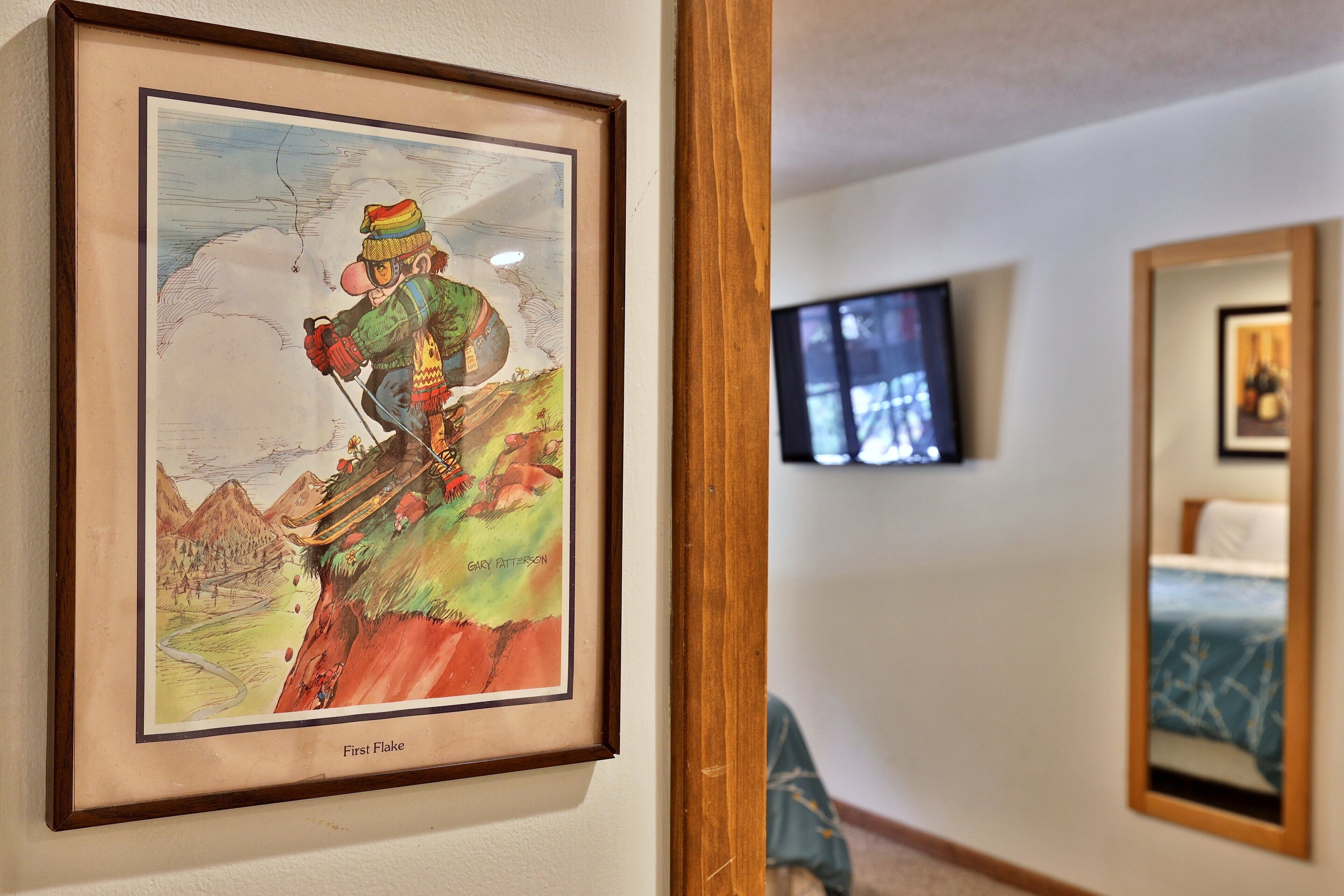 A framed artwork titled 'First Flake' by Gary Patterson, a mountain scene with a skier, mounted on a wall in a hotel room.