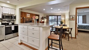 A modern kitchen with white cabinetry, a built-in microwave, and an oven. There is a dining area with a wooden table and chairs. - Snowplace Like Home at Mountain Green 1BR/1BA (Killington)