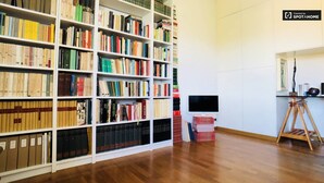 Eine Bibliothek mit weißen Bücherregalen, gefüllt mit Büchern, einem Fernseher auf einem Standfuß und einem Stativ auf dem Boden.