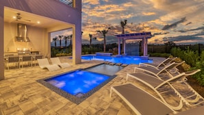 Un área de piscina moderna al aire libre con sillones de descanso, una pérgola y vistas a palmeras bajo un cielo al atardecer.