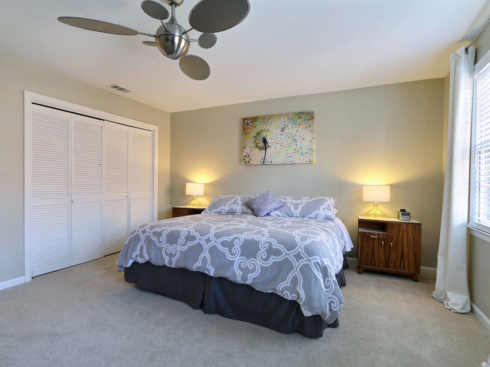 Une chambre à coucher comprenant un lit, deux tables de chevet, un ventilateur de plafond, un placard et une fenêtre avec des rideaux.