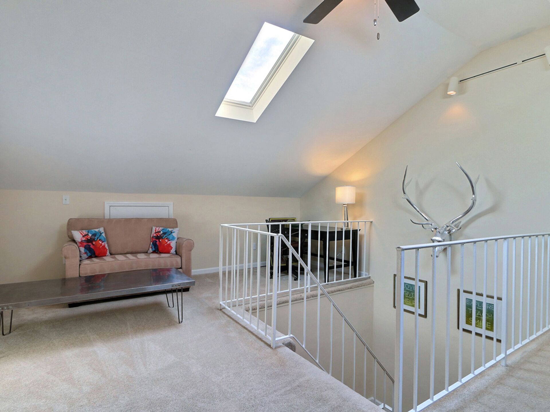 A spacious loft with a high ceiling, a large window, a sofa with colorful cushions, a metal coffee table, and a white railing with antlers.