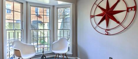 Una habitación con un ventanal grande, dos sillas blancas y un adorno de pared con una brújula roja.