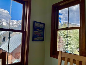 Ein Zimmer mit zwei Fenstern, einem gerahmten Gemälde an der Wand und Blick auf Berge und Bäume.
