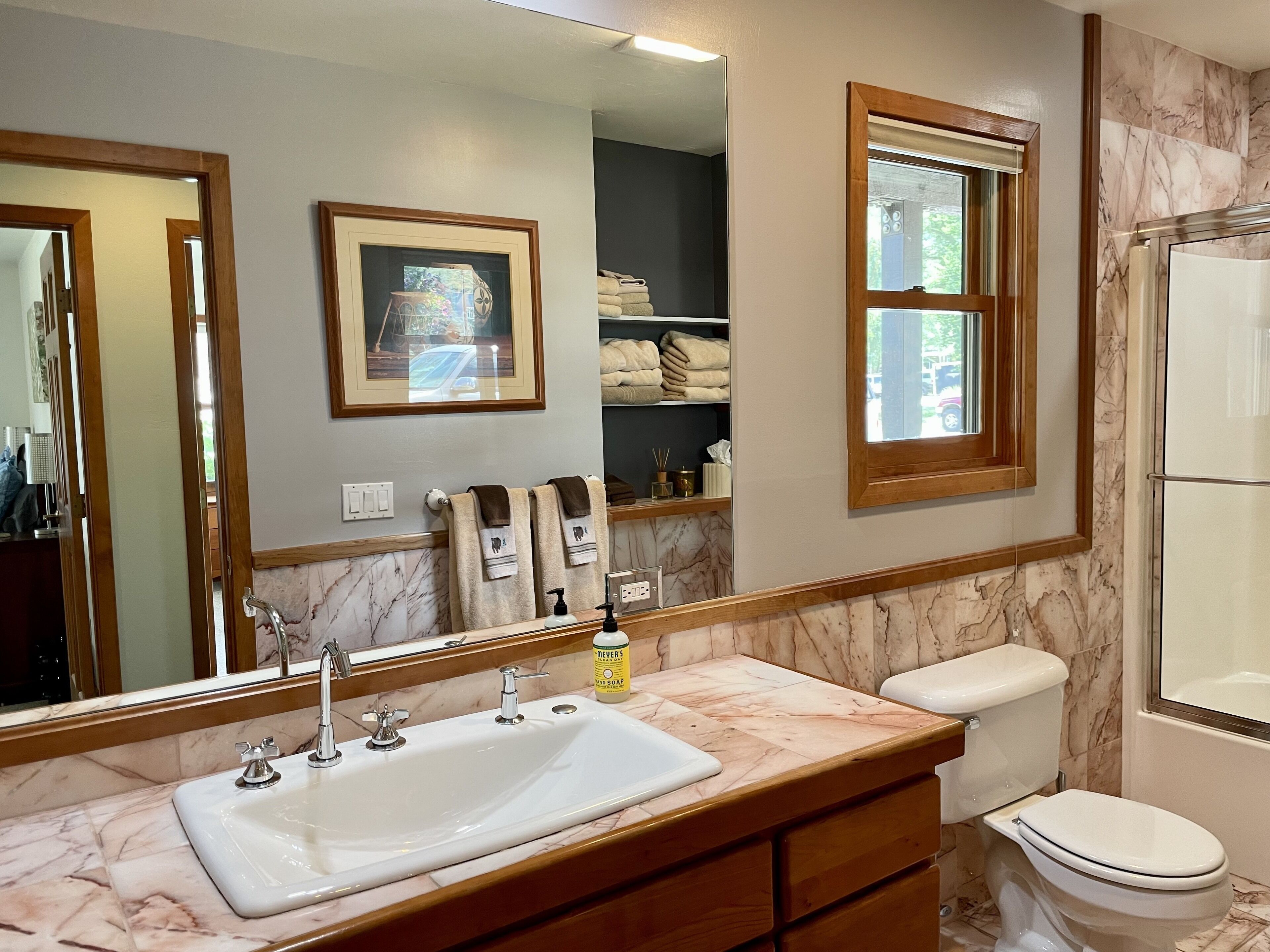 A bathroom with a marble sink, a toilet, a shower, a framed picture on the wall, and a window.