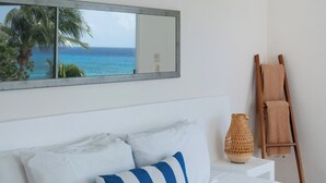 Un dormitorio con una cama, almohadas, una escalera de madera, una cesta tejida y vista al océano por una ventana.