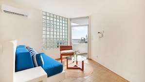 Una sala de estar moderna con un sofá blanco, cojines azules, una mesa de centro de vidrio, una silla de madera y vistas al exterior a través de una puerta de cristal.