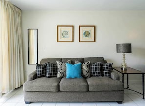 Ein graues Sofa mit gemusterten Kissen, ein gerahmtes Bild an der Wand, eine Lampe auf einem Beistelltisch und ein Vorhang am Fenster.