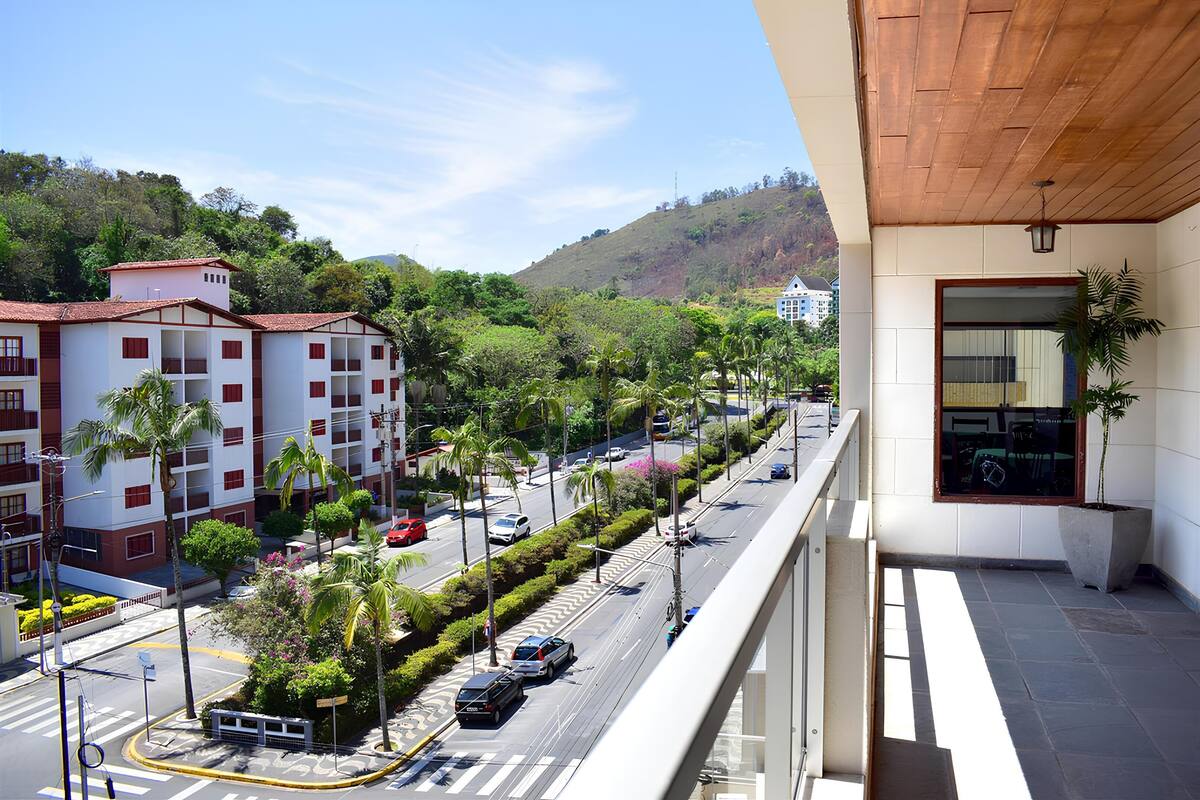Un hôtel à plusieurs étages doté de balcons, donnant sur une rue animée avec des voitures et de la verdure, et un paysage vallonné en arrière-plan.