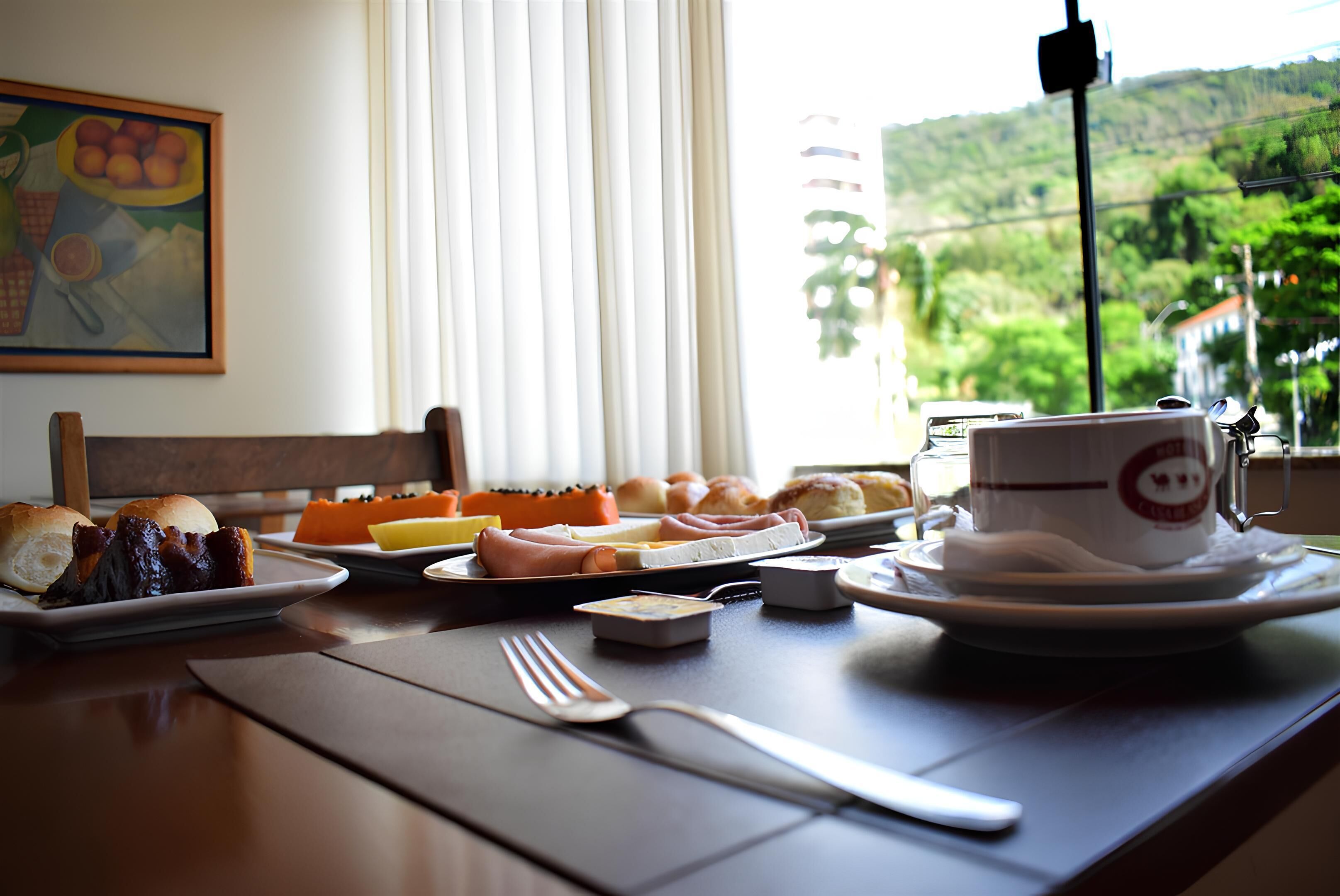 A breakfast table with various food items, a cup, and a view of greenery outside.