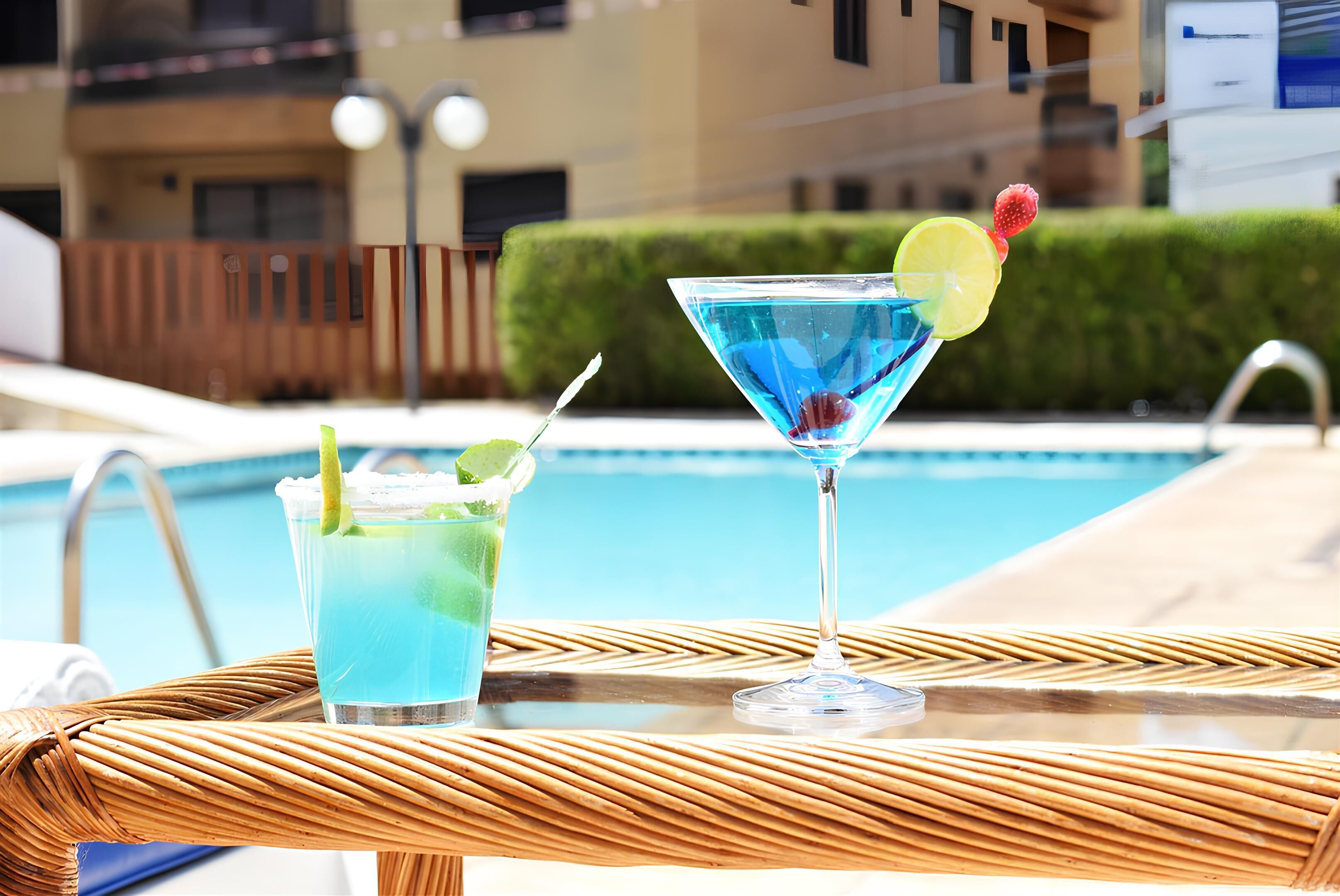 Two cocktails on a wicker table by a poolside.